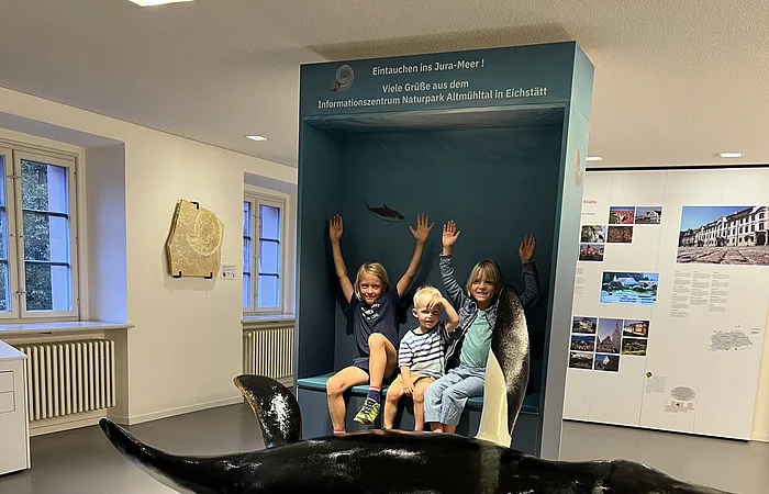 Drei Kinder sitzen vor einer blauen Museumsvitrine mit Text, davor ein großes Modell eines urzeitlichen Meeresreptils.