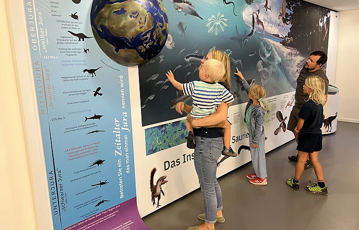 Familie betrachtet Museumsausstellung mit Dinosaurier- und Meereslebewesen-Wandbild und Erdkugel-Modell.