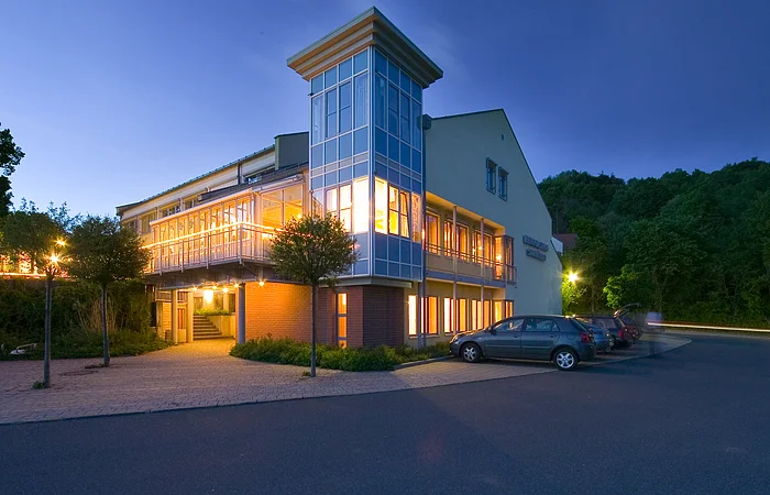 Beleuchtetes modernes Gebäude mit Parkplätzen und Bäumen bei Abenddämmerung unter klarem Himmel.