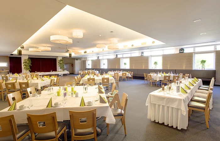 Großer heller Veranstaltungsraum mit eingedeckten Tischen und Holzstühlen, vorbereitet für ein Bankett.