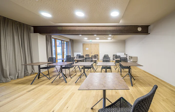 Leerer Schulungsraum mit Holzfußboden, mehreren Tischen und schwarzen Stühlen, Deckenleuchten und Vorhängen.