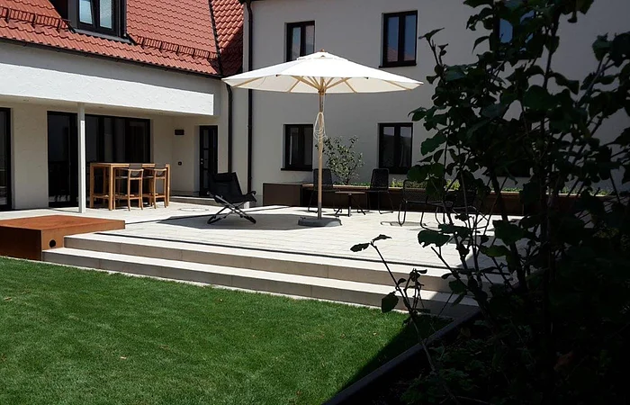 Terrasse mit Sonnenschirm, Stühlen und Tisch vor einem weißen Haus mit roten Dachziegeln und grünem Rasen.