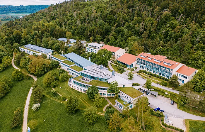 Luftaufnahme eines Gebäudekomplexes mit grünen Dächern am Waldrand und umliegenden Wiesen und Wegen.