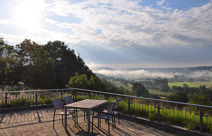 Terrasse mit Tisch und Stühlen, Blick auf neblige Landschaft mit Bäumen und Feldern bei bewölktem Himmel.