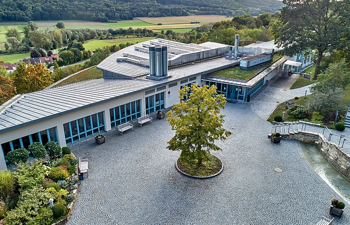 Luftaufnahme eines modernen Gebäudes mit großem gepflastertem Innenhof und Baum in der Mitte, umgeben von Grünflächen.
