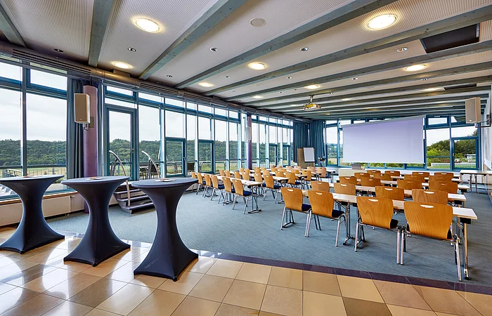 Konferenzraum mit Reihen von Tischen und Stühlen, drei Stehtische vorne und großen Fenstern mit Blick ins Grüne.