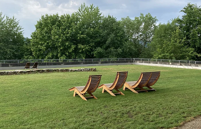 Vier hölzerne Liegestühle auf einer grünen Wiese vor Bäumen und bewölktem Himmel.
