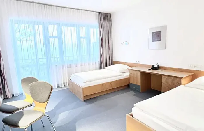 Hotelzimmer mit zwei Einzelbetten, Schreibtisch, zwei Stühlen und großen Fenstern mit Gardinen.