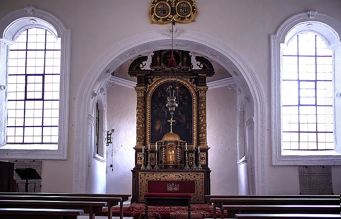 Innenraum einer Kirche mit Altar, zwei großen Fenstern und Holzbänken auf beiden Seiten
