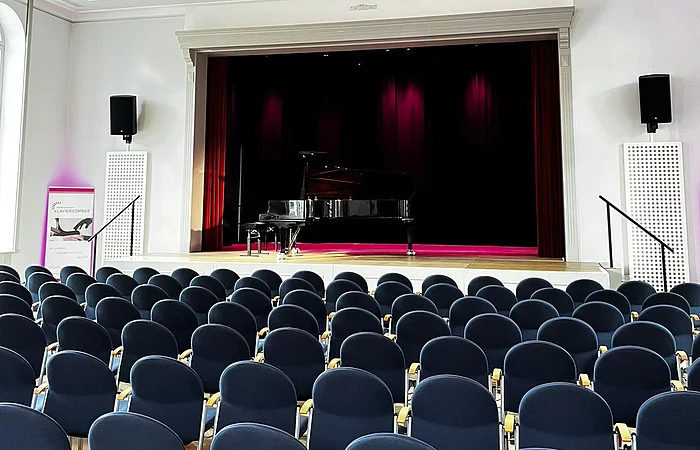 Leerer Konzertsaal mit Reihen blauer Stühle und einem Flügel auf der Bühne unter einer Deckenleuchte.