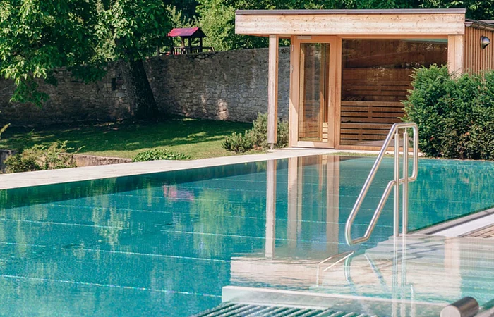Außenpool mit Edelstahlgeländer und Holzhütte vor großer Steinmauer und Bäumen im Garten.