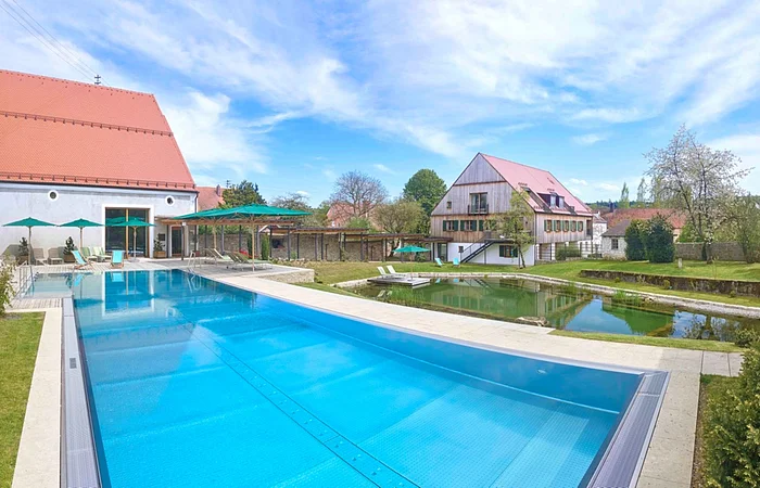 Außenbereich mit großem Schwimmbecken, Liegestühlen und Gebäuden mit roten Dächern bei blauem Himmel.