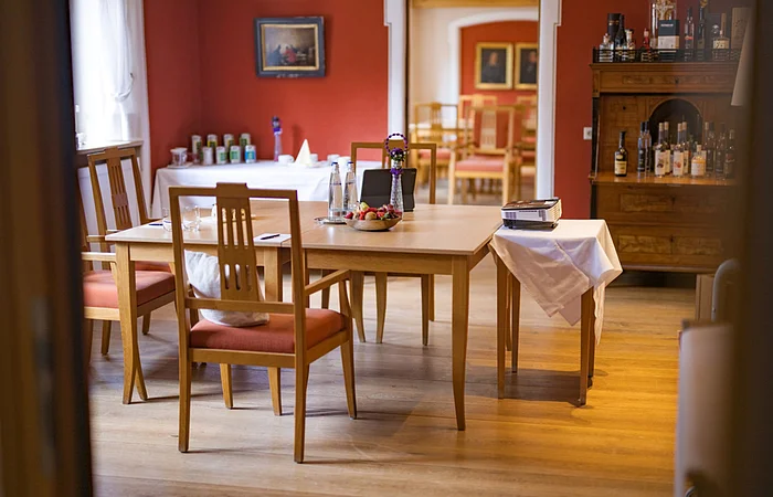Holzstühle und Tisch in einem Raum mit roter Wand, darauf Wasserflaschen, Obstschale und ein Beamer.