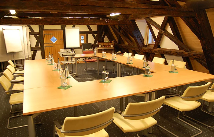 Konferenzraum mit U-förmigen Tischen, Stühlen, Wasserflaschen und Flipcharts unter Holzbalken.