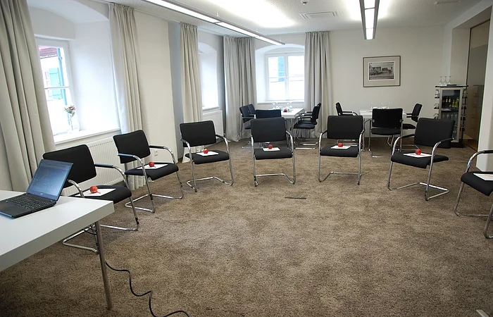 Konferenzraum mit Stühlen im Halbkreis, auf jedem Stuhl ein Apfel und ein Zettel, Laptop auf Tisch links.