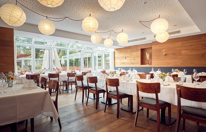 Restaurant mit gedeckten Tischen, Holzstühlen, großen Fenstern und runden Papierlampen an der Decke.