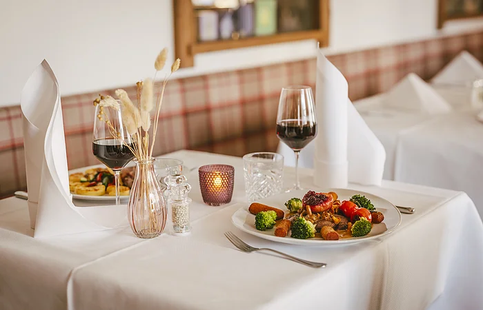 Tisch mit weißer Tischdecke, Teller mit Essen, zwei Weingläser, Vase mit Trockenblumen und Kerze in Restaurant.