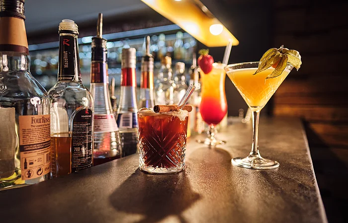 Bar mit verschiedenen Spirituosenflaschen und drei Cocktails auf der Theke, darunter ein brauner Drink mit Zimtstange.