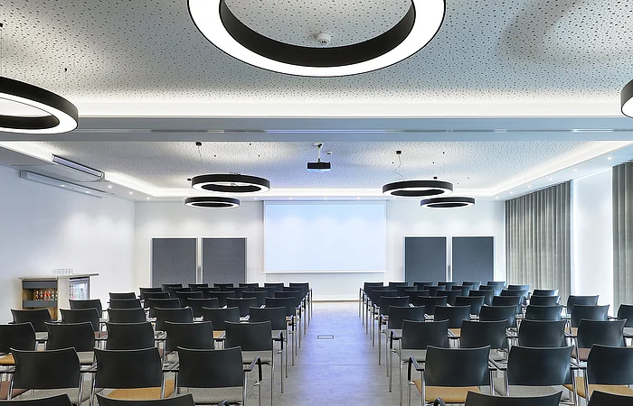 Konferenzraum mit Reihen schwarzer Stühle, Leinwand und runden Deckenleuchten in modernem Design.