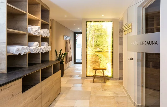 Flur mit Holzregalen und weißen Handtüchern, Tür mit Aufschrift „Kräutersauna“ und beleuchtetes Naturbild.