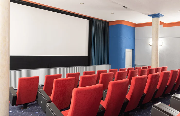 Leerer Kinosaal mit roten Sitzen, großer Leinwand und blauen und orangen Wänden.