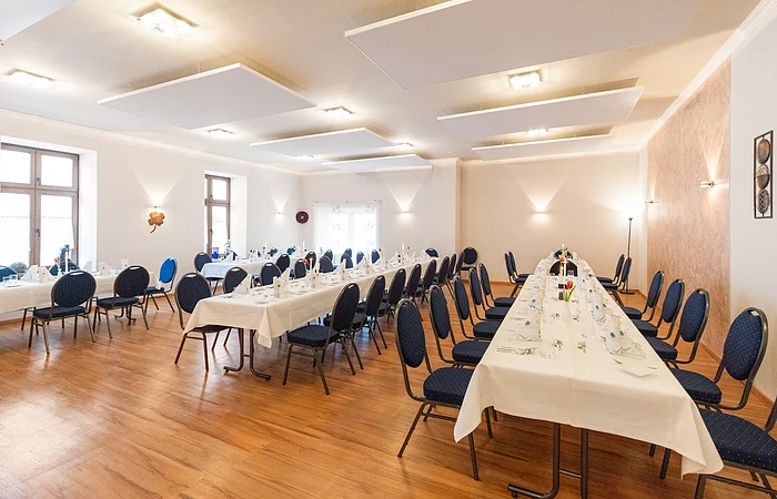 Großer heller Raum mit zwei langen gedeckten Tischen und blauen Stühlen auf Holzboden.