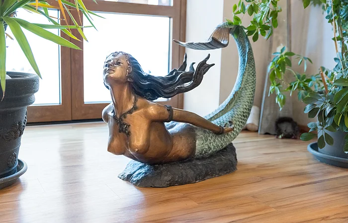 Bronzefarbene Meerjungfrauenskulptur mit grünem Fischschwanz auf Holzboden zwischen Pflanzen vor Fenster.