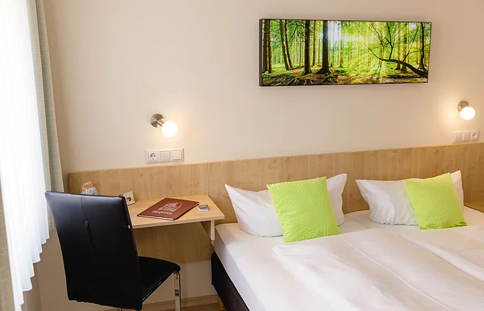 Hotelzimmer mit zwei Betten, grünen Kissen, Schreibtisch, schwarzem Stuhl und Waldbild an der Wand.