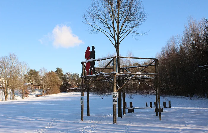 Holzplattform mit zwei Figuren, Schnee bedeckt, umgeben von Bäumen unter blauem Himmel