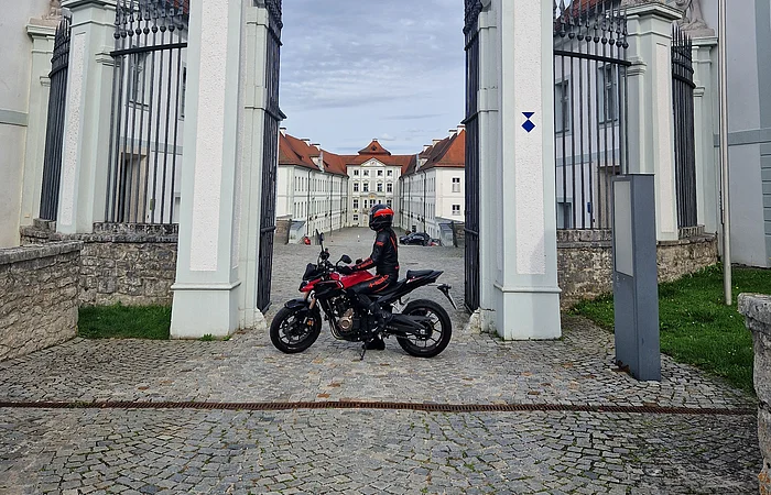 Motorradfahrer in schwarzer und roter Schutzkleidung vor Tor mit Hirschstatuen und historischem Gebäude im Hintergrund.