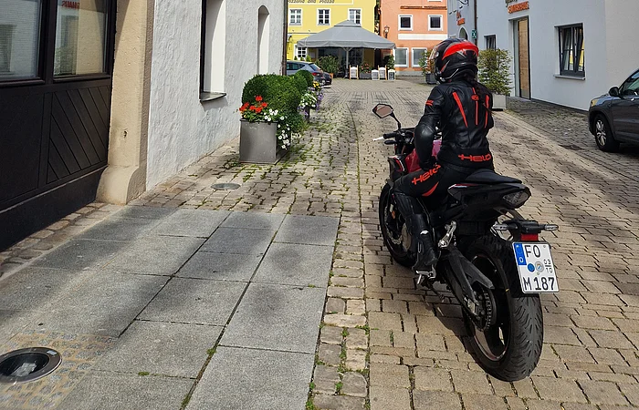 Motorradfahrer in schwarzer Schutzkleidung auf Kopfsteinpflasterstraße mit bunten Lampions und farbigen Häusern.