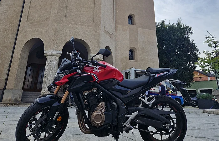 Rotes Motorrad steht vor einer Kirche mit zwei Türmen und einem Uhrenturm bei bewölktem Himmel.