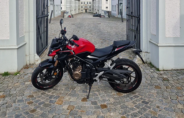 Rotes Motorrad steht auf gepflastertem Hof zwischen geöffneten schwarzen Toren vor weißen Gebäuden mit roten Dächern.