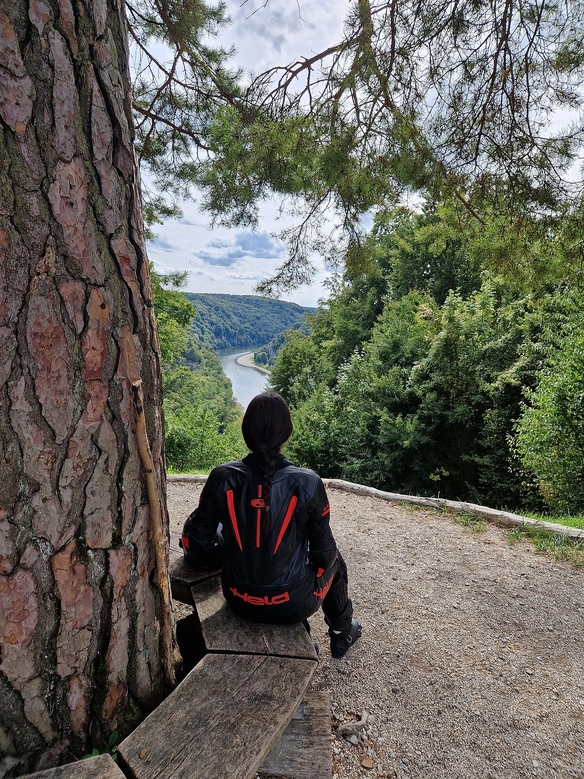 Person mit schwarzer Motorradjacke sitzt auf Holzbänken und blickt auf Fluss und bewaldete Hügel.