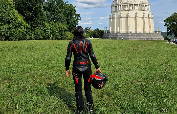 Person in schwarzer Motorradkleidung mit Helm in der Hand steht auf Wiese vor rundem Gebäude mit Säulen und Statuen.