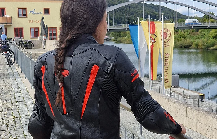 Person mit geflochtenem Haar und schwarzer Motorradjacke mit roten Akzenten lehnt an Geländer am Flussufer mit Brücke und Gebäuden im Hintergrund.