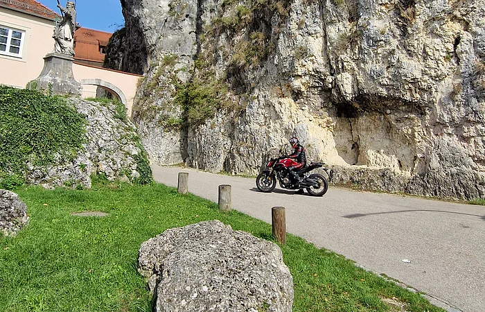 Motorradfahrer auf Straße vor steiler Felswand, links Statue und Gebäude mit rotem Dach.
