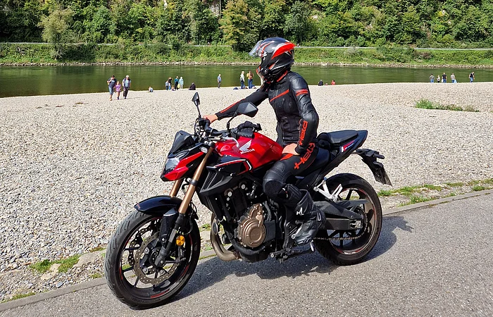 Person in schwarzer Motorradkleidung und Helm auf rotem Motorrad auf Weg am Flussufer mit Wald im Hintergrund