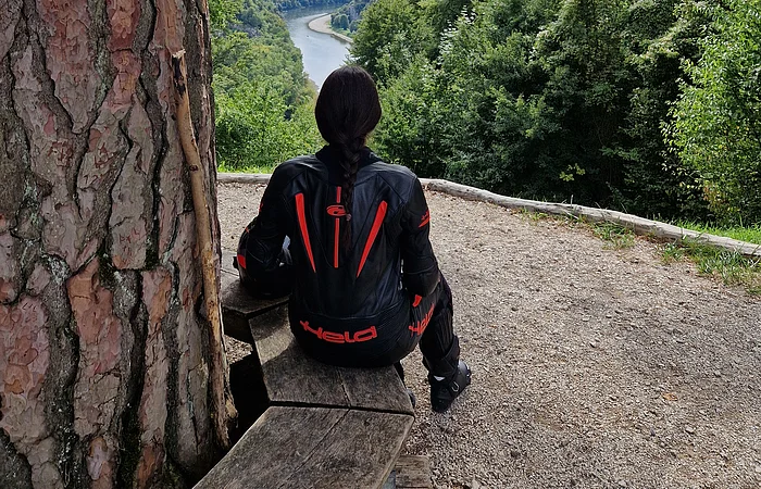 Person mit schwarzer Motorradjacke sitzt auf Holzbänken und blickt auf Fluss und bewaldete Hügel.