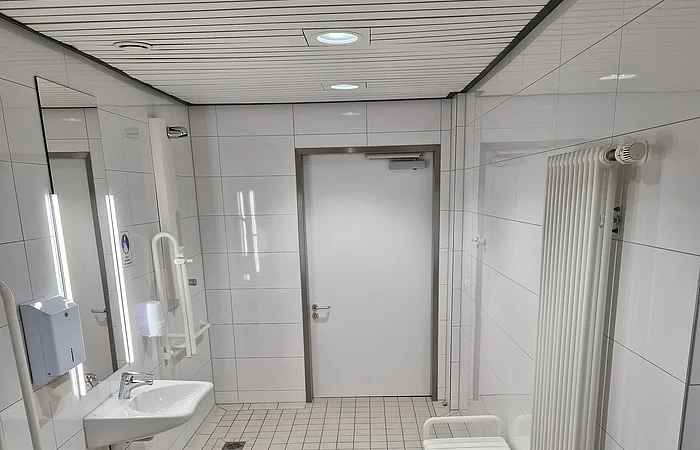 Barrierefreies Badezimmer mit Toilette, Waschbecken, Haltegriffen, Spiegel, Heizung und Sitzgelegenheit, alles in weiß/weiß gefließt.