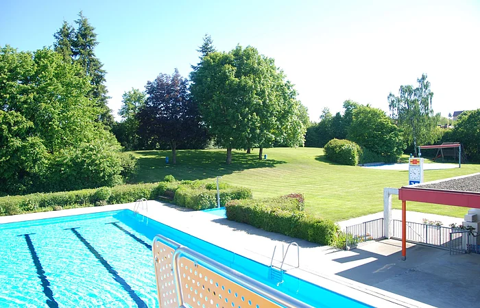 Außenbereich eines Schwimmbads mit Becken, Liegewiese, Bäumen und Uhr an einem sonnigen Tag.