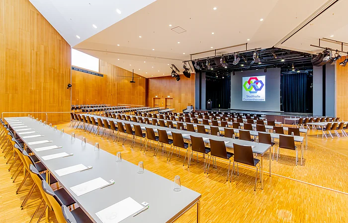 Großer Konferenzraum mit Reihen von Tischen und Stühlen, Bühne mit Rednerpult und Leinwand im Hintergrund.