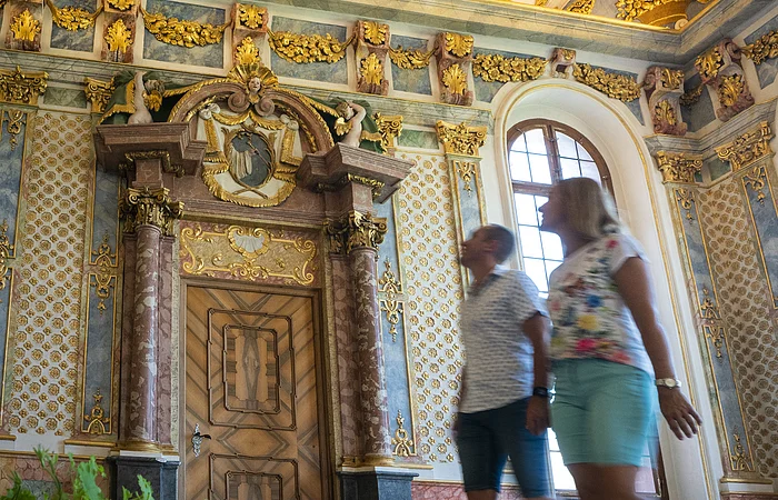 Zwei Personen gehen in einem prunkvoll verzierten Raum mit goldenen Stuckelementen und bemalter Decke.