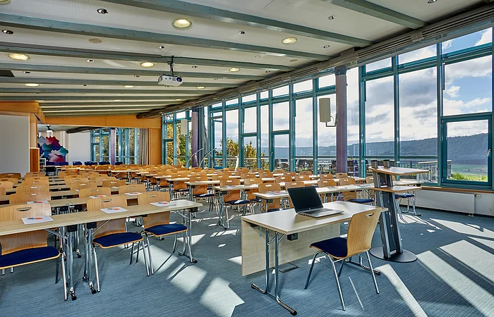 Großer Seminarraum mit Reihen von Tischen und Stühlen, Laptop auf vorderem Tisch, große Fenster mit Blick ins Freie.