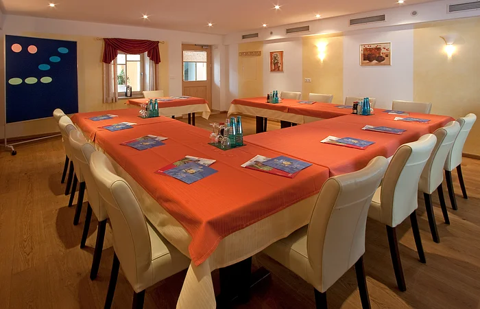 Konferenzraum mit U-förmiger Tischanordnung, orangen Tischdecken und weißen Stühlen, mit Flipchart und Fenstern.