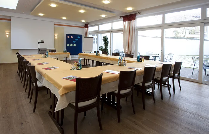 Konferenzraum mit U-förmiger Tischanordnung, Stühlen, Flipchart, Projektor und großen Fenstern mit Blick nach draußen.