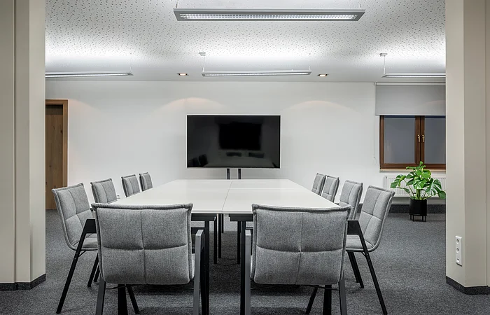 Konferenzraum mit weißem Tisch, zwölf grauen Stühlen, großem Bildschirm und Pflanze am Fenster.