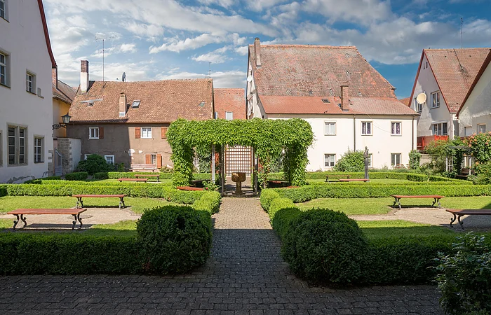 Innenhof mit gepflegtem Garten, Bänken, Hecken und alten Häusern mit roten Ziegeldächern im Hintergrund.