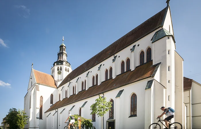 Weißes Kirchgebäude mit Turm und zwei Radfahrern im Vordergrund bei blauem Himmel.