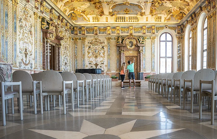 Prächtig verzierter Saal mit goldenen Stuckverzierungen, Sternmuster auf dem Boden und zwei Personen in der Mitte.